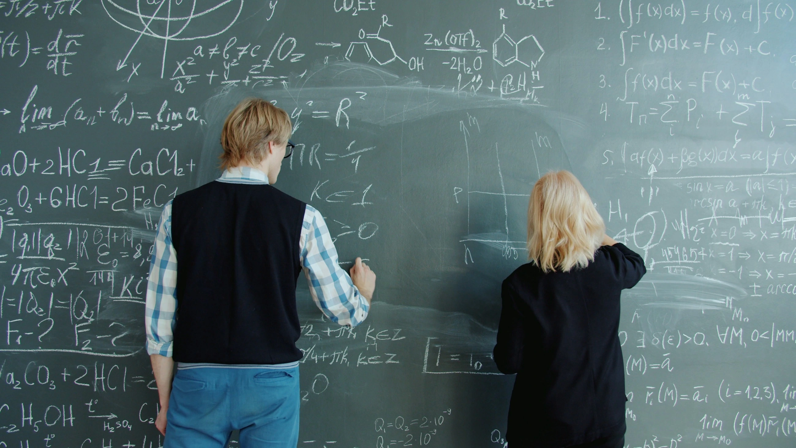 Two people are working with mathematical formulas on a blackboard.
