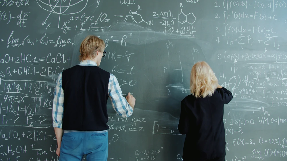 Two people are working with mathematical formulas on a blackboard.
